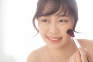 skönhetsporträtt ung asiatisk kvinna leende med ansiktet tittar spegeln applicera makeup med borste kinden i sovrummet, vacker flicka håller rouge, hudvård och kosmetiska koncept. foto