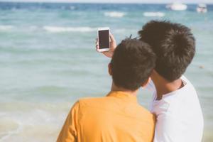 bakifrån gay porträtt ungt par leende tar en selfie foto tillsammans med smart mobiltelefon på stranden, lgbt homosexuell älskare i semestern till sjöss, två man kommer att resa, semester koncept.