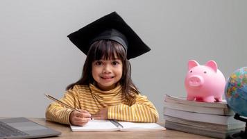 asiatisk liten flicka som bär en examensmössa med en rosa spargris, sparar pengar, investerar i framtiden, foto