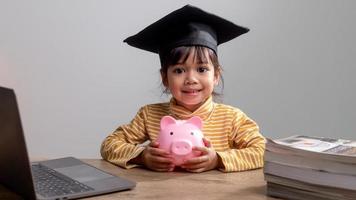 asiatisk liten flicka som bär en examensmössa med en rosa spargris, sparar pengar, investerar i framtiden, foto
