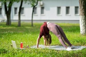 ung flicka med online-träning utomhus med hjälp av bärbar dator. pilates eller yoga videolektion på internet. glad leende tjej tränar pilateslektion online i trädgården utomhus under karantän. foto