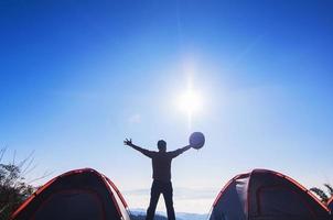 man glad camping står och höjer sin hand som håller sin hatt på berget på morgonen under sitt vandringsläger nära två små tält med solmoln och blå himmel bakgrund, thailand foto