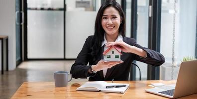 miniatyr hus i händerna på en asiatisk kvinna fastighetsmäklare bostadslån som arbetar på kontoret. tittar på kameran. foto