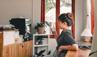 vacker asiatisk kvinna sätter sig vid bardisken i ett fönsterkafé och håller en kopp kaffe och ler avslappnat på ett kafé foto