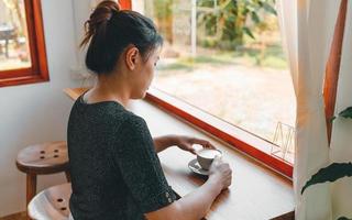 vacker asiatisk kvinna sätter sig vid bardisken i ett fönsterkafé och håller en kopp kaffe och ler avslappnat på ett kafé foto