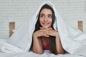 drömmande ung kvinna tittar ut ur en filt medan liggande i sängen hemma foto