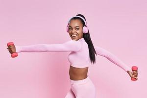 glad afrikansk kvinna i hörlurar tränar med hantlar mot rosa bakgrund foto