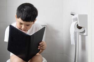 asiatisk pojke sitter på toalettskålen medan du läser bok - hälsoproblem koncept foto