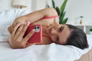 vacker ung kvinna som använder smart telefon medan du ligger i sängen hemma foto