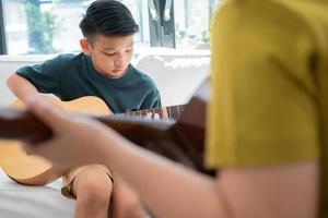 Asiatisk pojke spelar gitarr med pappa i vardagsrummet för att han lärde honom son spela gitarr, känner sig uppskattad och uppmuntrad. begreppet en lycklig familj, lärande och rolig livsstil, älskar familjeband foto