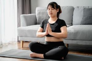 asiatisk knubbig kvinna sitter på golvet i vardagsrummet praktik yoga lektion. kvinna med meditera träningsklass. foto