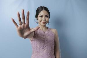 ung asiatisk kvinna som bär modern kebaya över blå isolerad bakgrund gör stoppskylt med handflatan. foto