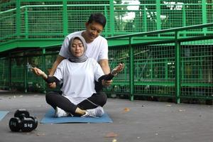 ung asiatisk man hjälper sin fru att göra meditation i parken. hälsosam livsstil par koncept. foto