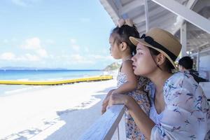 glad dotter och mamma sitter på resturant nära havet och tittar på vacker utsikt över den tropiska stranden. människor tittar på himmel och blått hav. resa livsstil på familjens sommarsemester med barn foto