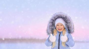 liten skrattande flicka med tummen upp på den snöiga vintersolnedgången foto