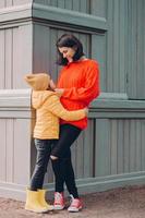 vertikal bild av attraktiv omtänksam mamma tittar på ögonen på sin dotter med kärlek, omfamning medan poserar utanför, bär trasiga jeans, tröja och sneakers, le positivt. familjeförhållande koncept foto