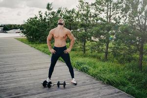 frisk muskulös man står med naken överkropp, höjer huvudet och andas djupt, bär sportkläder, tränar med skivstänger, poserar utomhus. bodybuilding, aktiv livsstil, fitness koncept foto