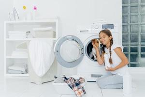 foto av attraktiv tjej som klappar stamtavla i tvättmaskin, håller i tvättmedel, går och laddar tvättmaskin, upptagen med tvätt och hushållssysslor, tvättar kläder hemma, poserar inomhus.