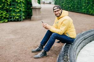 upprymd man med skägg och charmigt ansikte klädd i trendiga kläder som vilar utomhus och är alltid i kontakt med sin smartphone. förtjusande hipster man i gul anorak isolerad över grön bakgrund foto