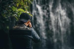 närbild av baksidan fotograf i casual stil bär jacka och hatt använder modern kamerafotografering med suddigt vattenfall. foto