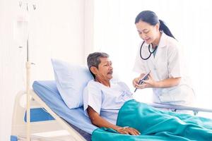 asiatisk kvinna professionell läkare med urklipp som besöker, pratar och diagnostiserar den gamla manpatienten som ligger i patientens säng på sjukhusavdelningen foto