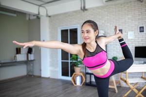 ung asiatisk kvinna tränar och gör yoga i den stående bågen som drar poserar hemma, hälsosamt koncept. foto