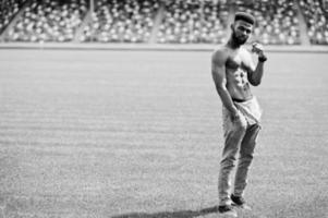 stilig sexig afrikansk amerikansk muskulös bar överkropp man på jeansoveraller poserade på grönt gräs på stadion fotbollsplan. fashionabla svart man porträtt. foto