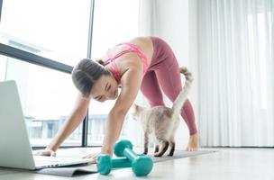 ung asiatisk vacker kvinna tränar kondition hemma med katt och tittar på onlinehandledning öva träning träningsworkshop på bärbar dator. foto