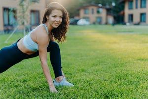 energisk kvinna i topp och leggings, gör stretchövningar på grön gräsmatta under solig dag utomhus, värmer upp innan konditionsträning. atletisk dam i sportkläder tycker om fitness. sport, rekreation foto