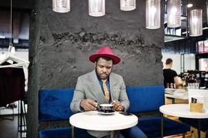 snygg afrikansk amerikansk man modell i grå jacka slips och röd hatt dricka kaffe på café. foto