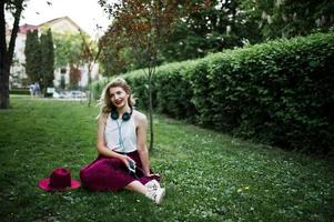 moderiktig och vacker blond modellflicka i snygg röd sammetsvelourkjol, vit blus och hatt, sittande på grönt gräs i parken med telefon och hörlurar. foto