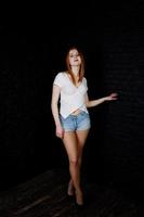 porträtt av en vacker rödhårig tjej i vit t-shirt och jeansshorts poserar i studion bredvid tegelväggen. foto