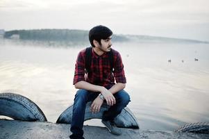 ung indisk student man på rutig skjorta och jeans med ryggsäck poserade på kvällen stad mot sjön. foto