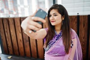 indisk hinduisk flicka på traditionell violett saree sitter vid kafébordet med mobiltelefonen vid händerna och gör selfie. foto