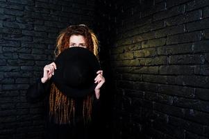 studiofotografering av flicka i svart med dreads och hatt på tegelbakgrund. foto