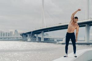 full längd skott av muskler aktiv man gör sportövningar utomhus poser nära floden bridge bär byxor och sneakers har vacker manlig överkropp uppnår bästa form. hälsosam livsstil koncept foto