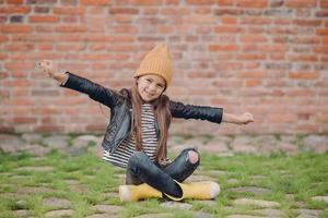 vacker liten flicka sitter i lotusställning, sträcker ut armarna, bär gul hatt, skinnjacka och riggade jeans, poserar över en tegelvägg i det fria, har ett positivt ansiktsuttryck. barn och roligt koncept foto