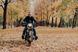 brutal professionell manlig motorcyklist cyklar, bär solglasögon, handskar och svart jacka, har åkt genom höstparken, vacker natur i bakgrunden med gula träd och fallna löv runt om foto