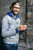 fantastisk man med blå ögon, skägg och brett leende bär keps, jeansskjorta och glasögon som håller smartphone isolerad över gammal vägg bakgrund. nöjd man i snygga kläder med mobiltelefon foto