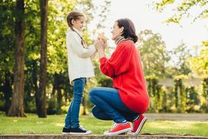 ung mamma i varm stickad röd tröja leker med sin lilla dotter i parken, ger henne löv, njut av soligt höstväder. tillgiven mamma och lilla barn spenderar tid tillsammans utomhus foto