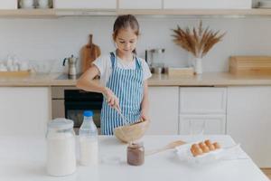 liten söt tjej i förkläde, blandar ingredienser, vispar med visp, använder ägg, mjölk, mjöl, provar nytt recept, står emot köksinredning, förbereder läckra kakor eller bageri, lär sig laga mat. foto