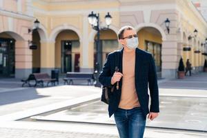 utomhusbild av allvarlig man går på stationen med ryggsäck, koncentrerad åt sidan, bär medicinsk mask för att förhindra virus, väntar på buss eller tåg, bryr sig om hälsan. coronavirus pandemi koncept. foto