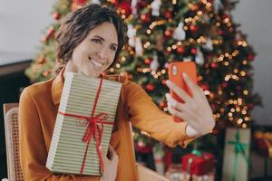 glad italiensk kvinna sitter nära vacker julgran hemma och tar selfie med present foto