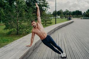 atletisk man står i sidoplankpose, höjer ena armen, poserar med bar överkropp, bär byxor och smyger. idrottsman övningar i parken på frisk luft, har perfekt passform muskler kropp. hälsosam livsstil foto