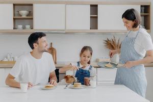 glad familj tid och frukost koncept. glad fru och mamma förbereder läckra pannkakor för familjemedlemmar, far, dotter och hund njuter av att äta och smaka efterrätt hemma, lägg till choklad foto
