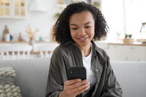 ung biracial flicka chattar eller postar på sociala nätverk, använder smartphone-appar, sitter på soffan hemma foto