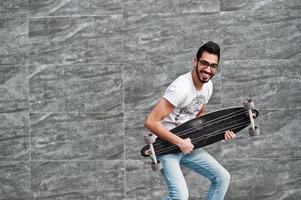 street style arabisk man i glasögon med longboard poserade mot grå vägg, som om han spelar gitarr. foto