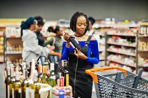 afrikansk kvinna väljer vin på snabbköpet mot hennes afro vänner med kundvagn. foto