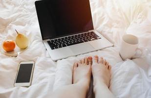 närbild kvinna fötter avkopplande på sängen med en bärbar dator, smartphone, te och frukt på vitt lakan foto