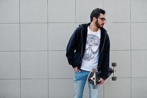 street style arabisk man i glasögon med longboard poserade mot grå vägg. foto